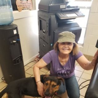 Woman kneeling with a dog, near a photocopier and water cooler. She's wearing a hat and purple shirt, smiling.