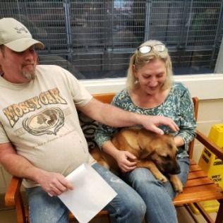 A couple pets a brown dog on a bench inside; man holds paper, woman smiles.