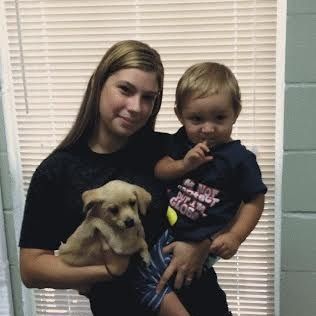 Woman holding a puppy and a child in front of blinds. The boy is wearing a blue shirt.