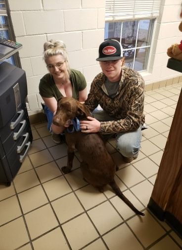 Two people kneel with a brown dog; one person pets the dog. The setting is indoors near a window.