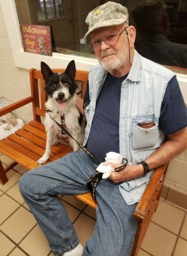 Man in denim vest sits on bench with black and white dog, inside.