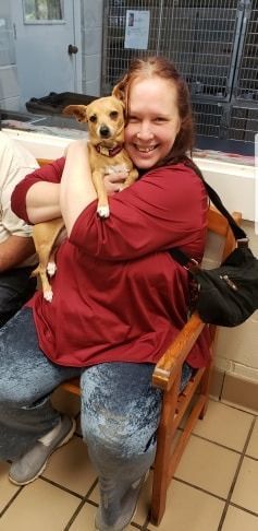 Woman sitting and holding a small brown dog in her arms; smiling. They are indoors by a cage.