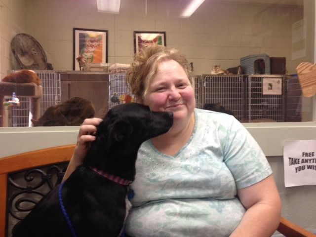 Woman smiles as black dog nuzzles her face inside a shelter.  Other animals and cages visible in background.