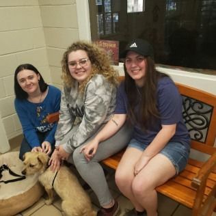 Three people seated with a dog on a bench. They smile while petting the dog. Indoors near a window.