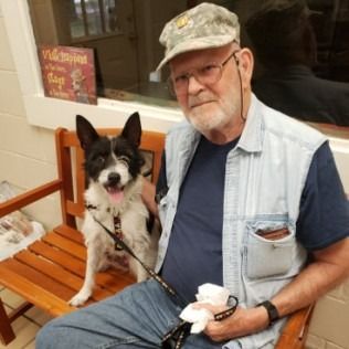 Man in cap and vest sits with black and white dog on a wooden bench indoors.