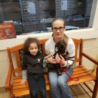 Woman and child sit on a bench, holding a small dog in a shelter.
