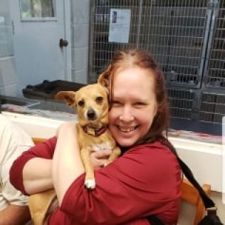 Woman in red shirt hugs a small tan dog at an animal shelter, both smiling.