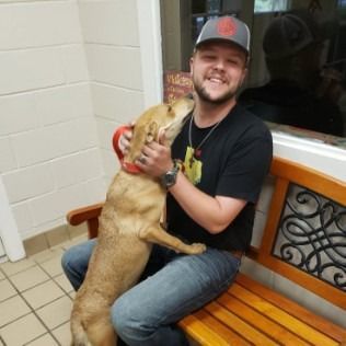 Man sitting on a bench is embracing a tan dog, indoors.
