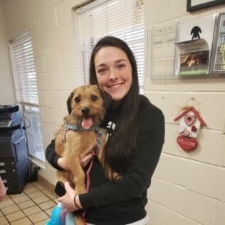 Woman smiling, holding a small brown dog indoors near a window. The dog has its tongue out.