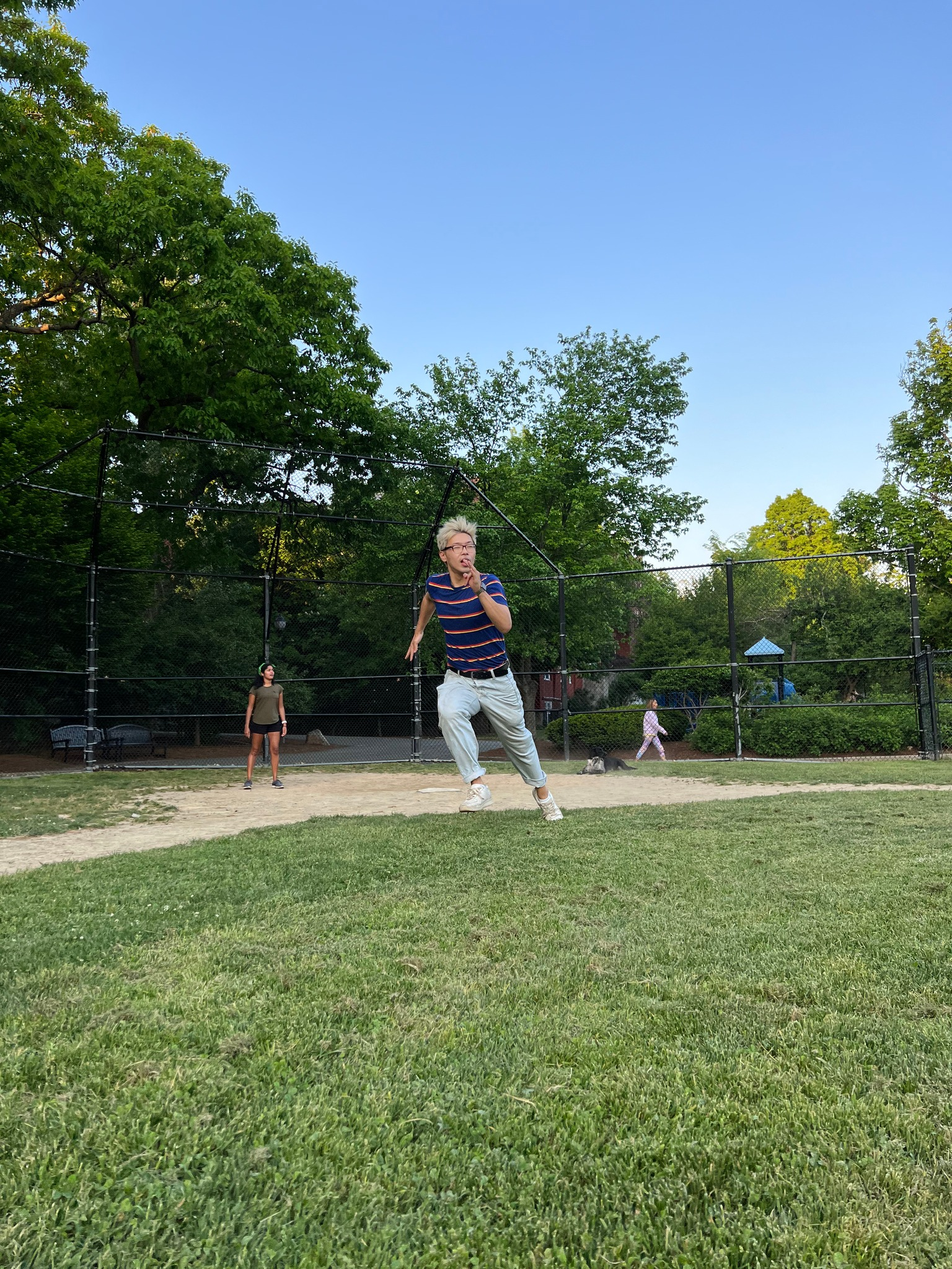 Blond-haired person runs on a grassy area near a black fence. Trees and a park in the background.