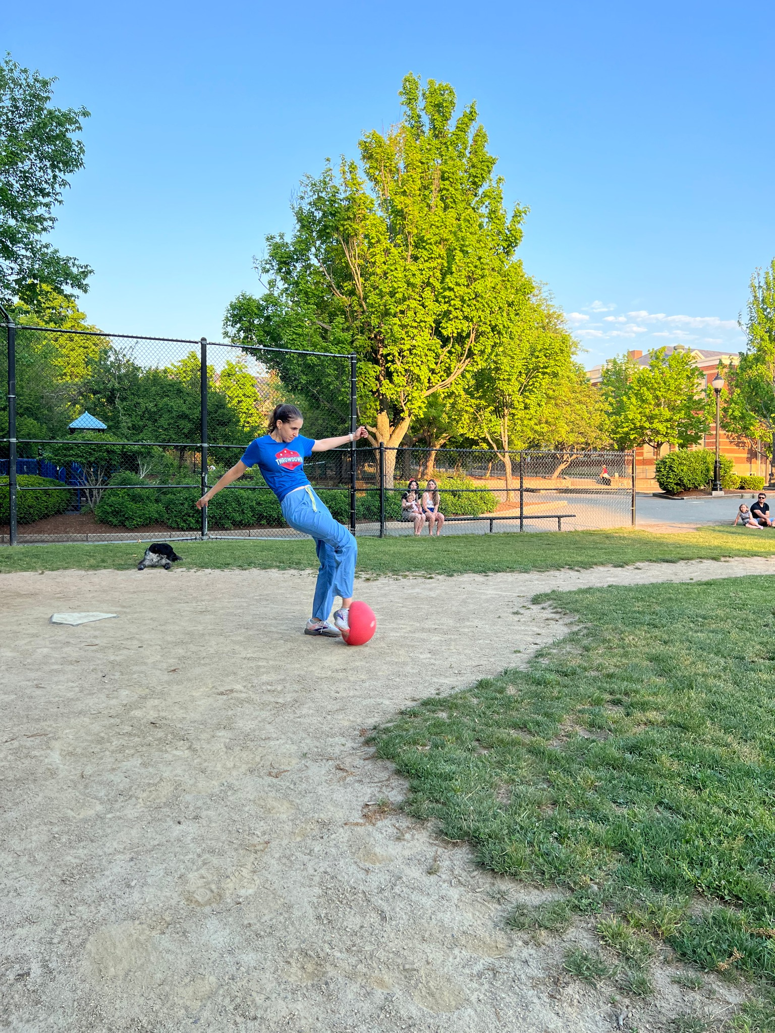 Person kicking a red ball in a park. They are wearing blue jeans and a blue shirt. The setting is outdoors.