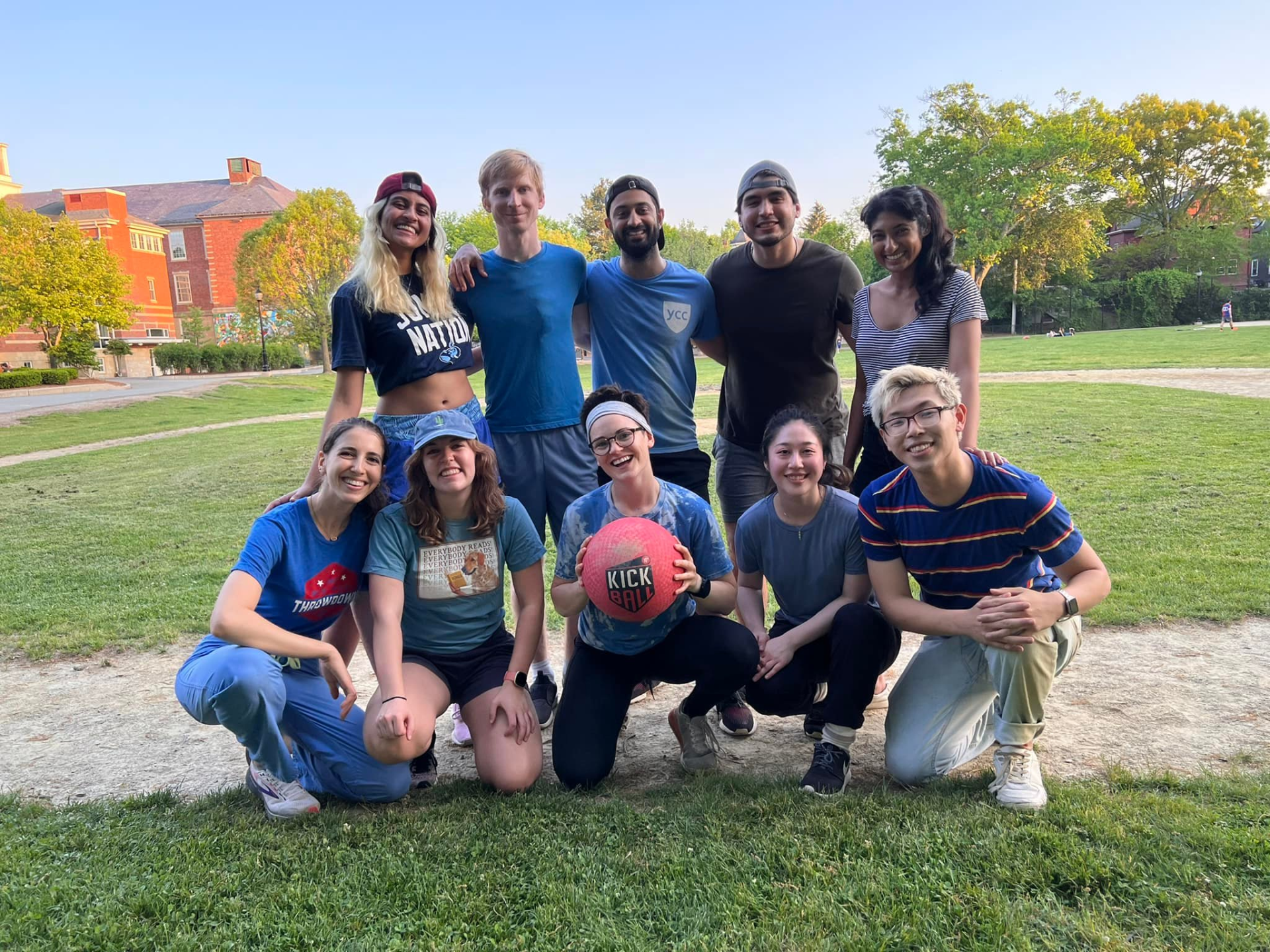 Group of people posing with a red ball on a grassy field; several are kneeling, others standing, smiling.
