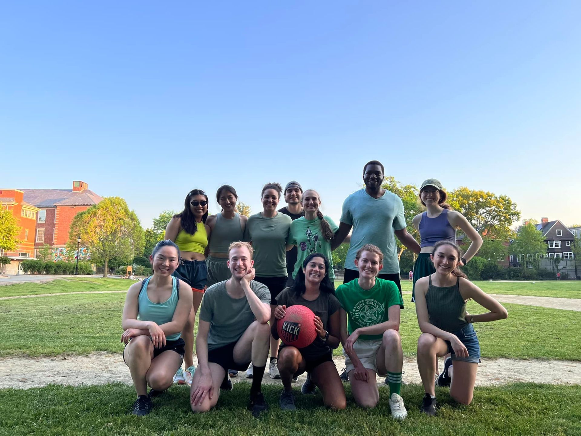 Group of people in green posing on a grassy field with a red ball; buildings and blue sky in the background.