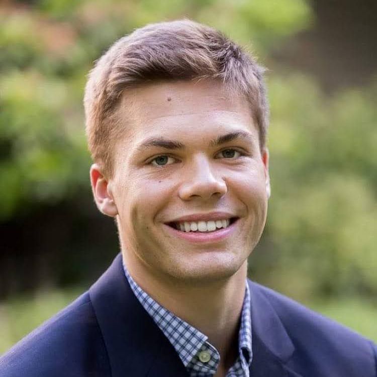 Man in a blue blazer smiles, checkered shirt underneath, outdoors, blurred green background.
