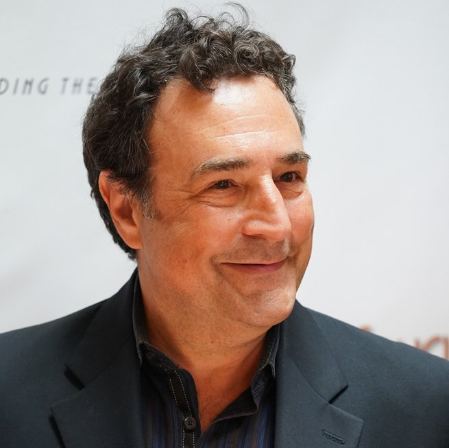 Man in a dark suit smiles, head slightly turned. Curly dark hair, neutral background.