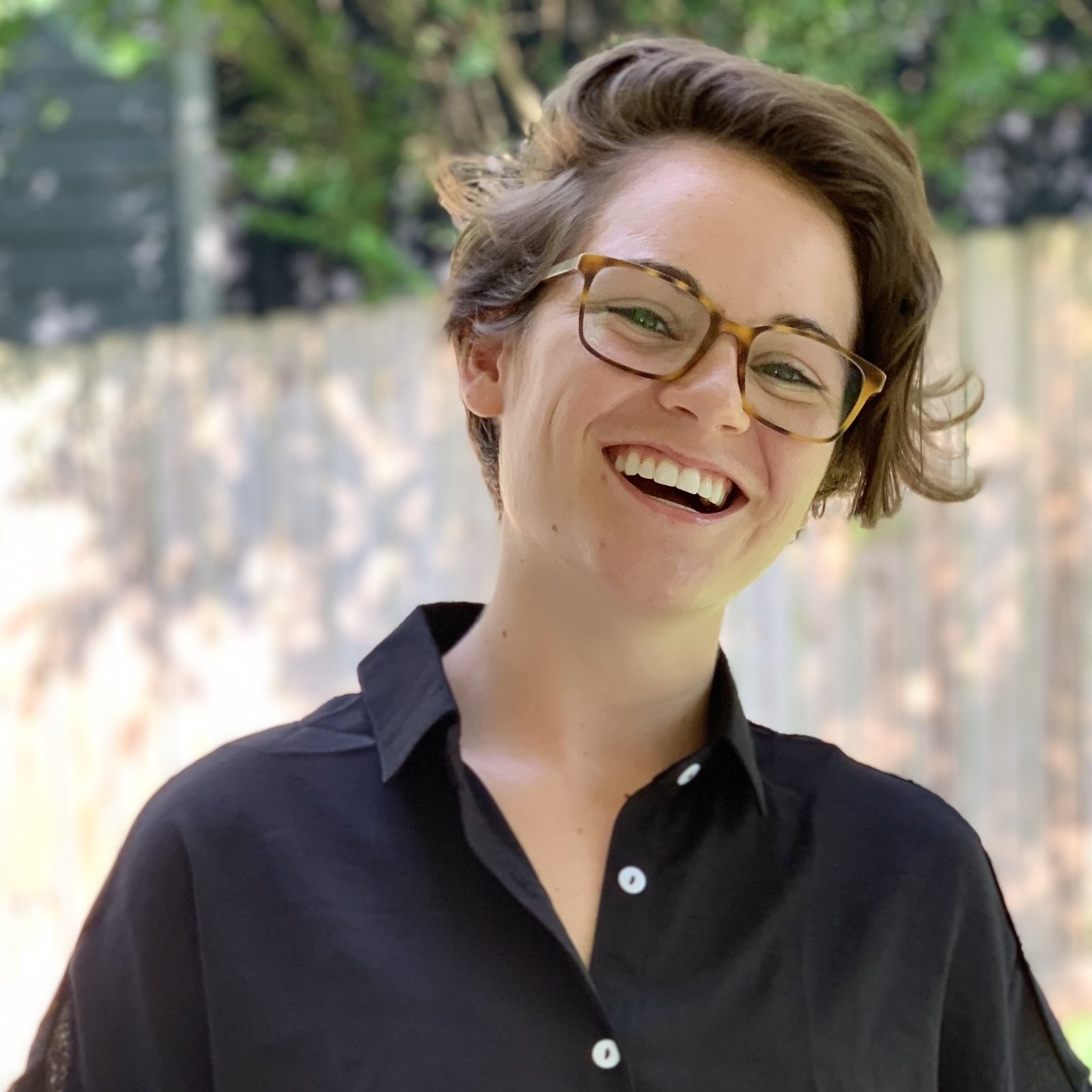 Woman with glasses smiles, wearing black shirt outdoors.