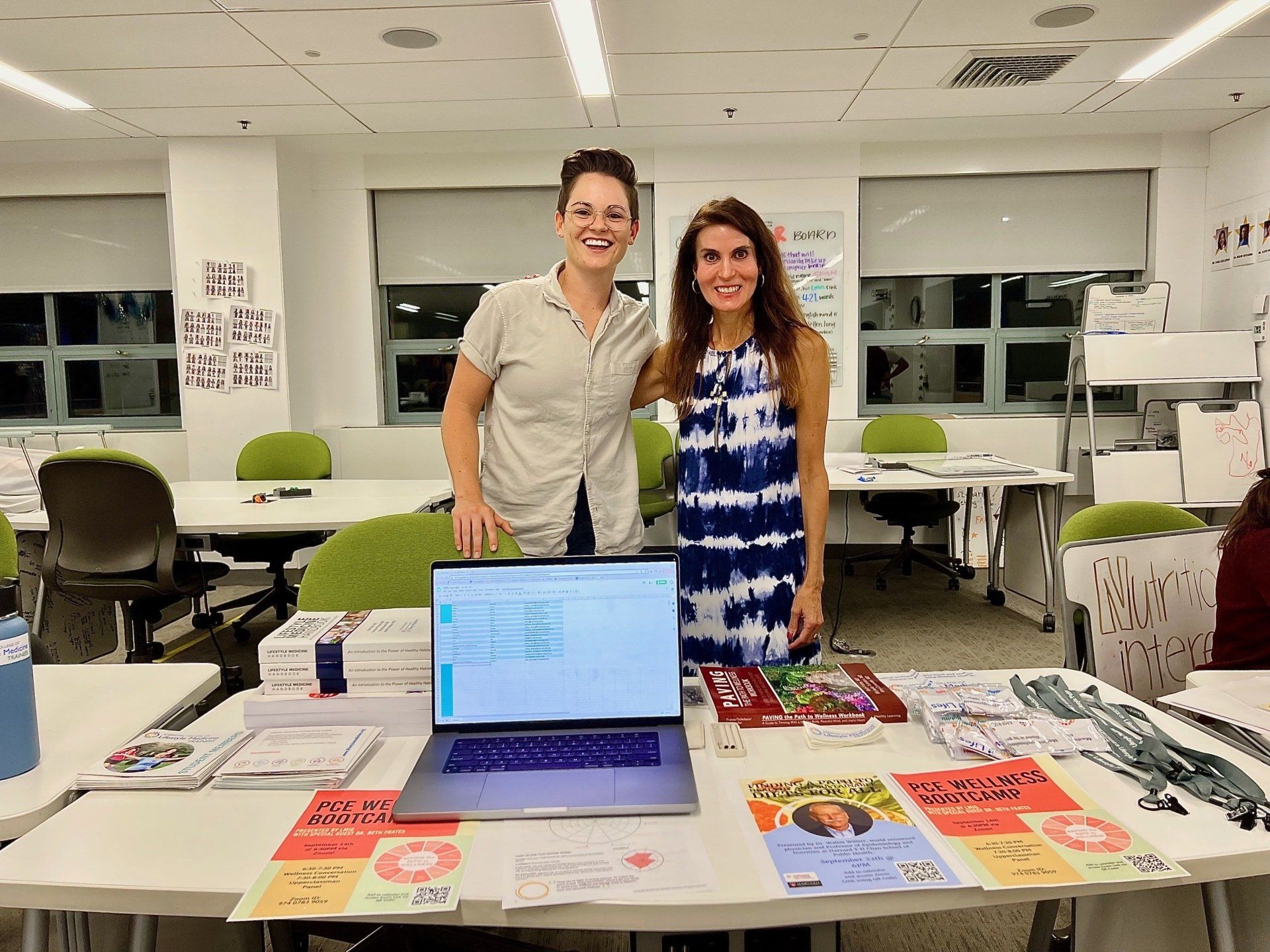 Two women smiling behind a table with laptop, books, and promotional materials in a brightly lit room.