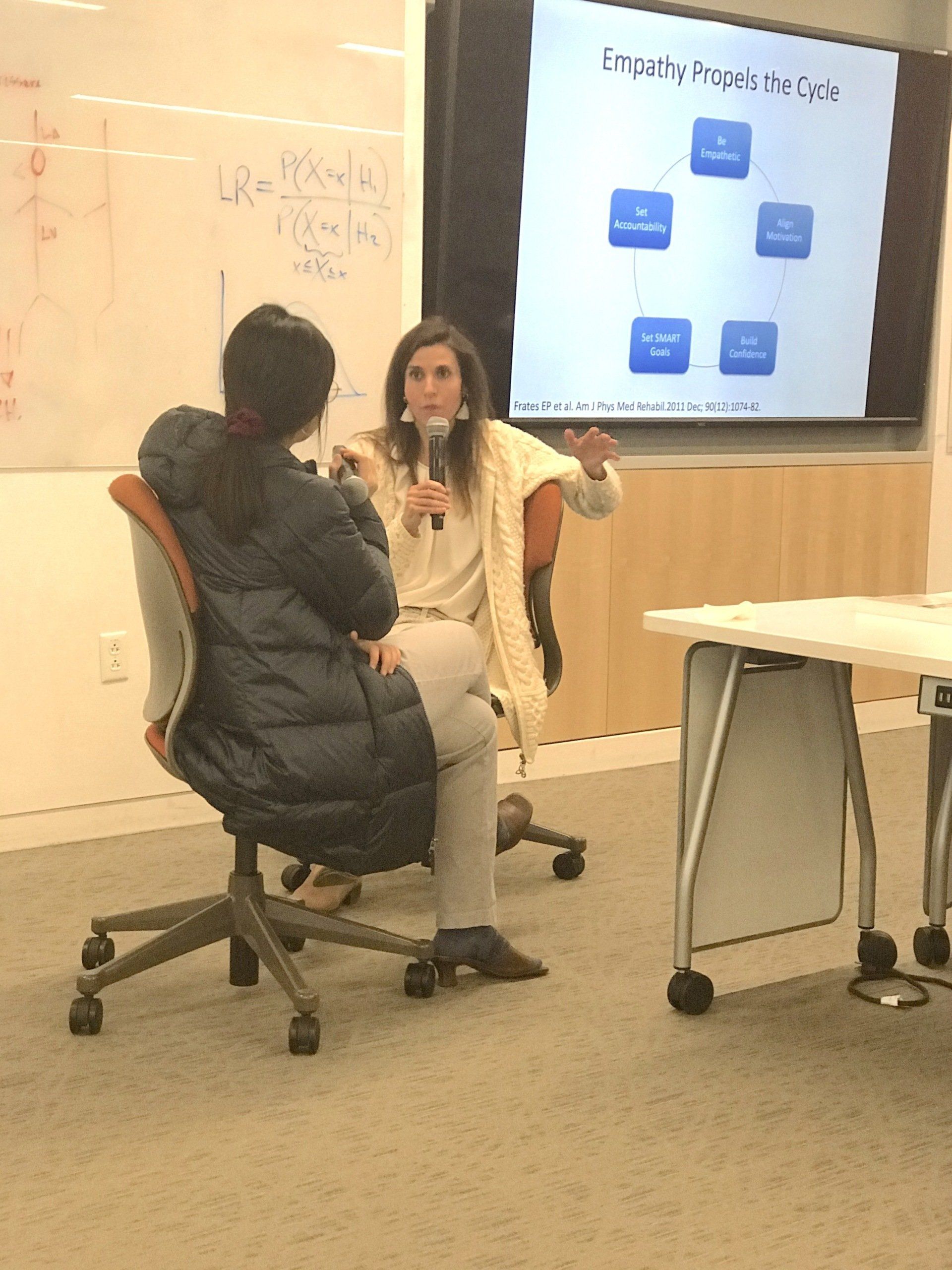 Woman speaking, gesturing, holding a microphone, facing a seated person. Presentation screen in background, whiteboard to the left.