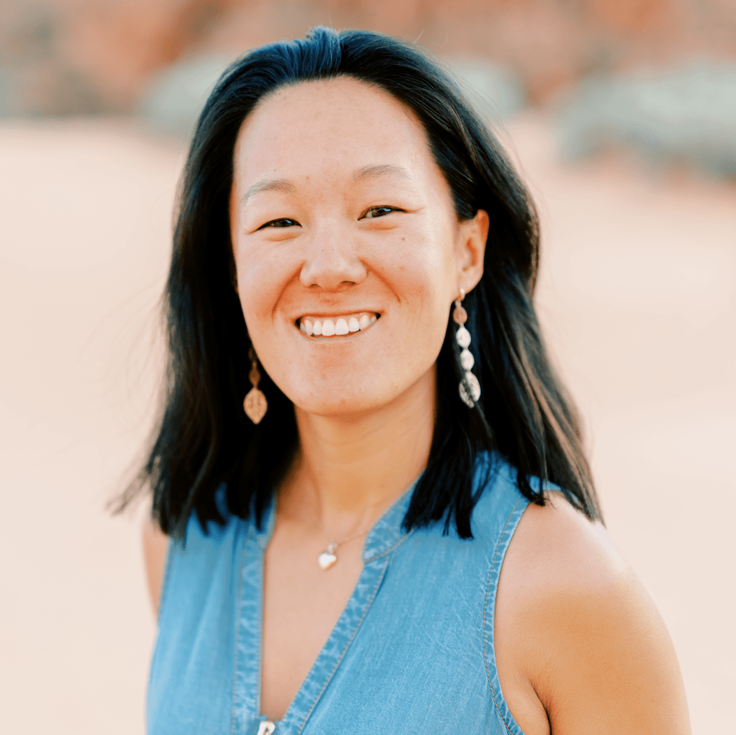 Woman with dark hair smiles, wearing blue dress, earrings, necklace, outdoors.