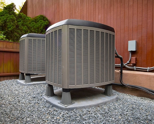 Two outdoor HVAC units on gravel beside a wooden building with pipes and a utility box.