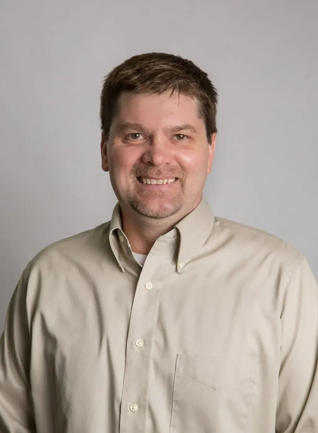 Man in a light-colored button-down shirt smiles, standing against a gray backdrop.