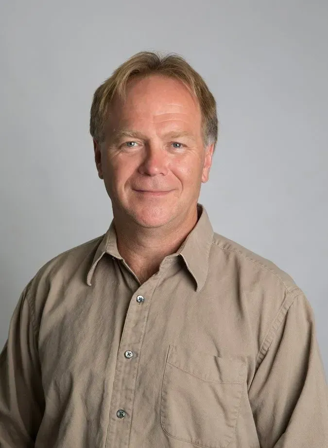 Man with short blonde hair and a tan button-down shirt smiles in front of a neutral grey background.