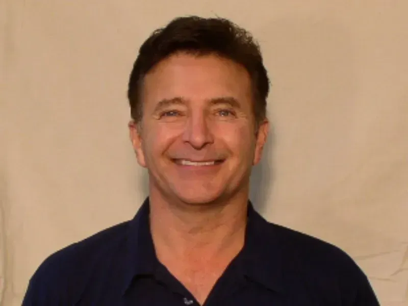 Man in blue shirt smiles directly at camera, neutral background.