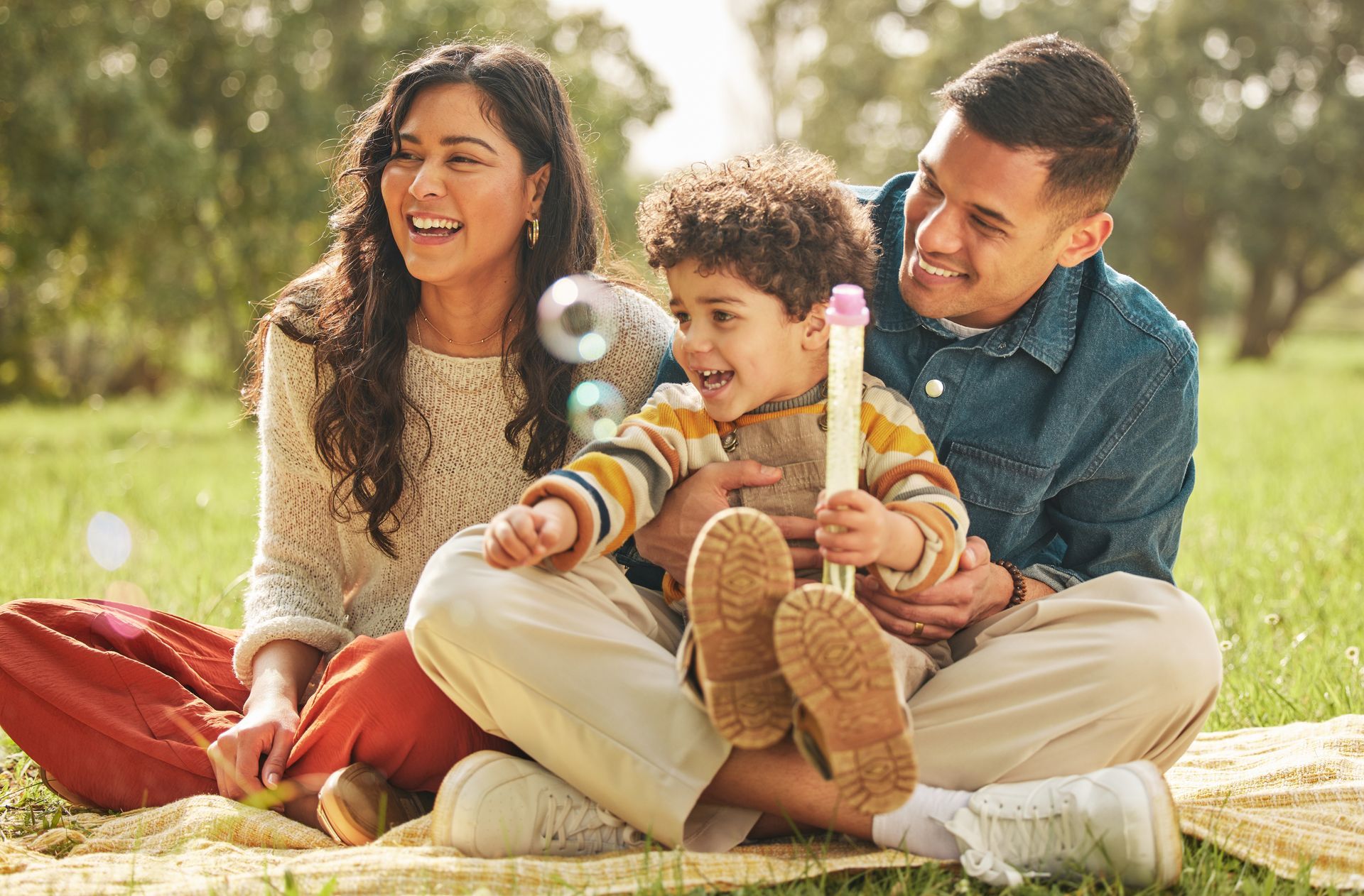 Family of three enjoying a park. Child plays with bubbles; parents smile, sitting on a blanket.