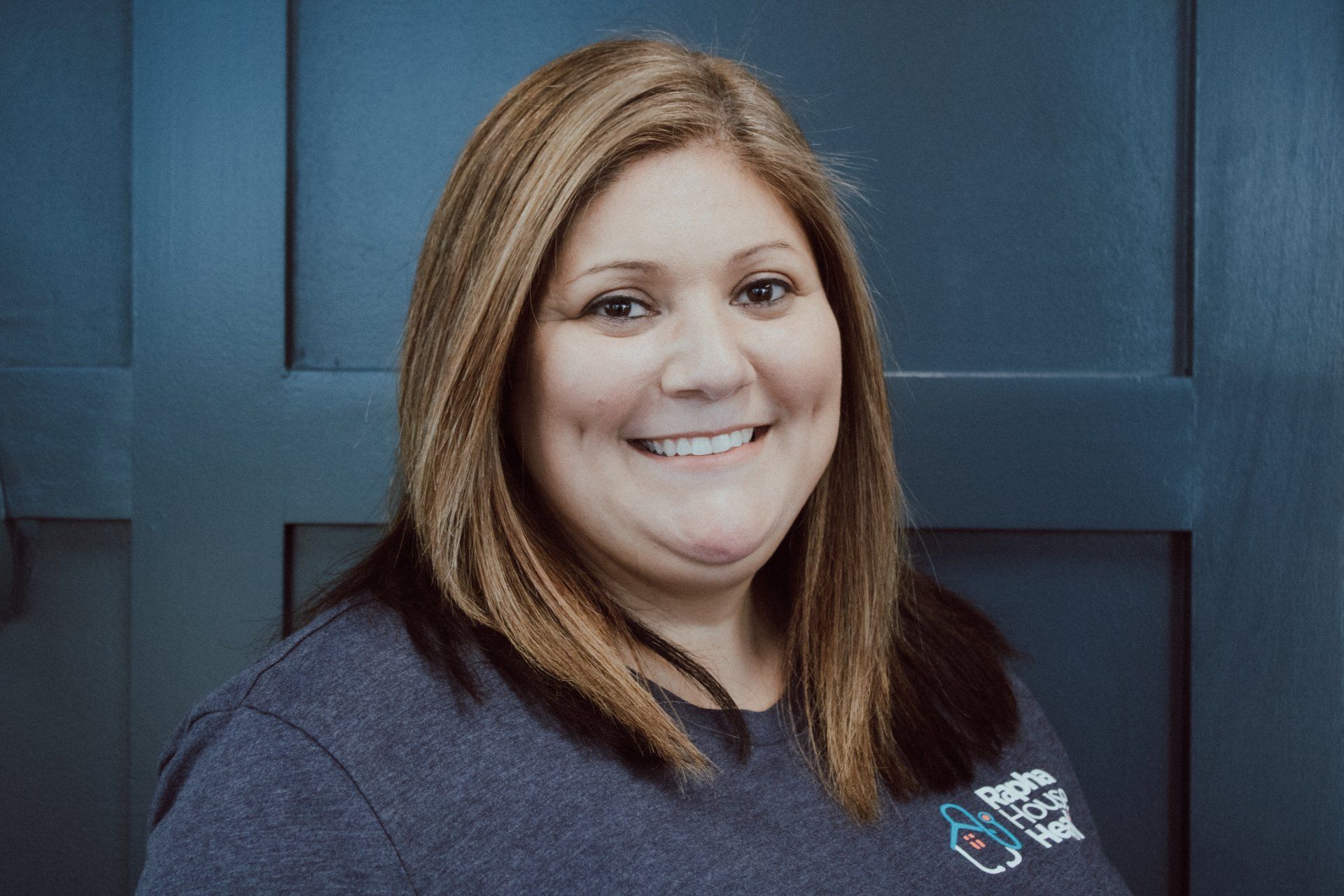 A woman in a gray shirt is smiling in front of a blue door.