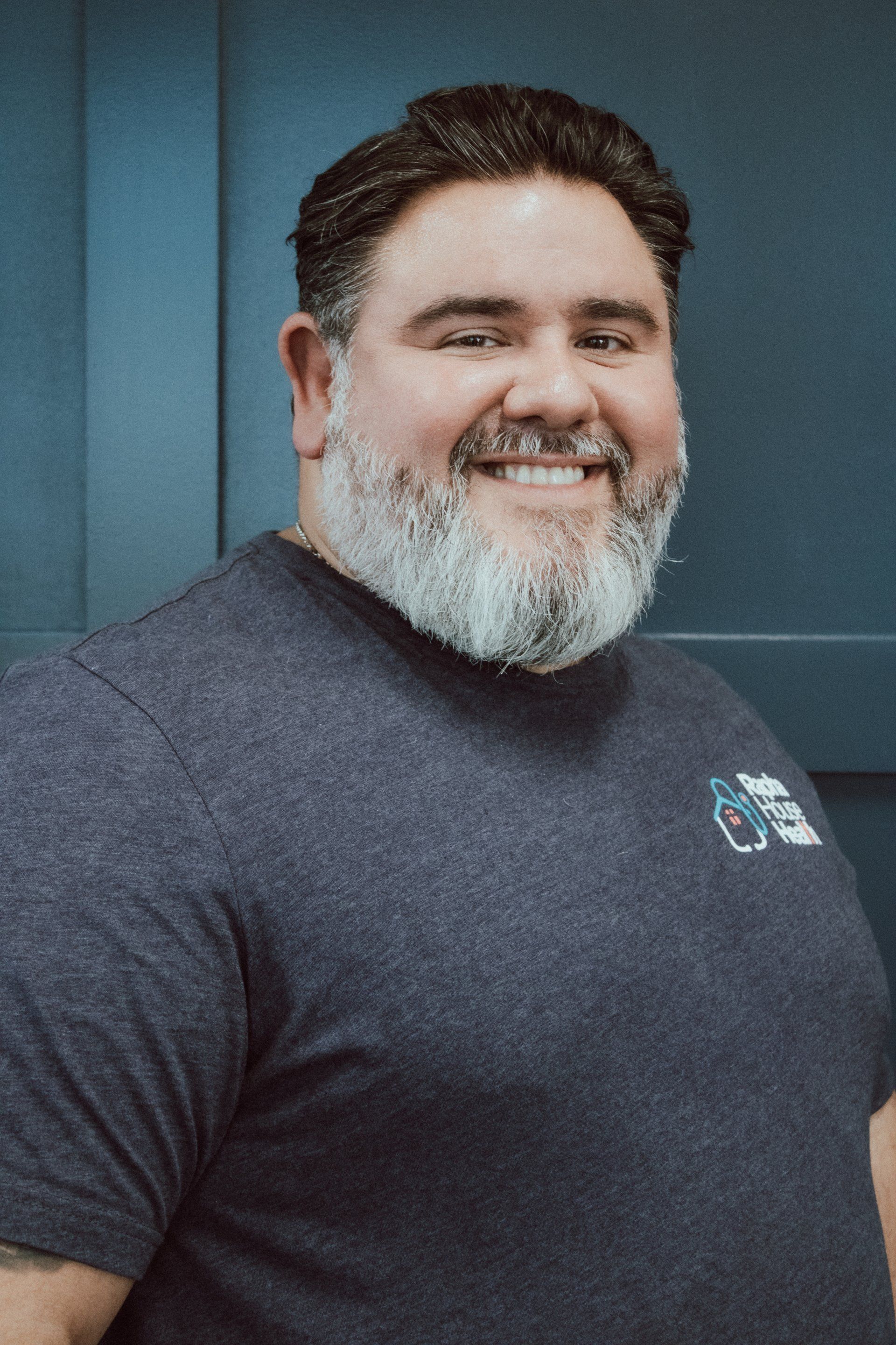 A man with a beard is wearing a gray t-shirt and smiling.