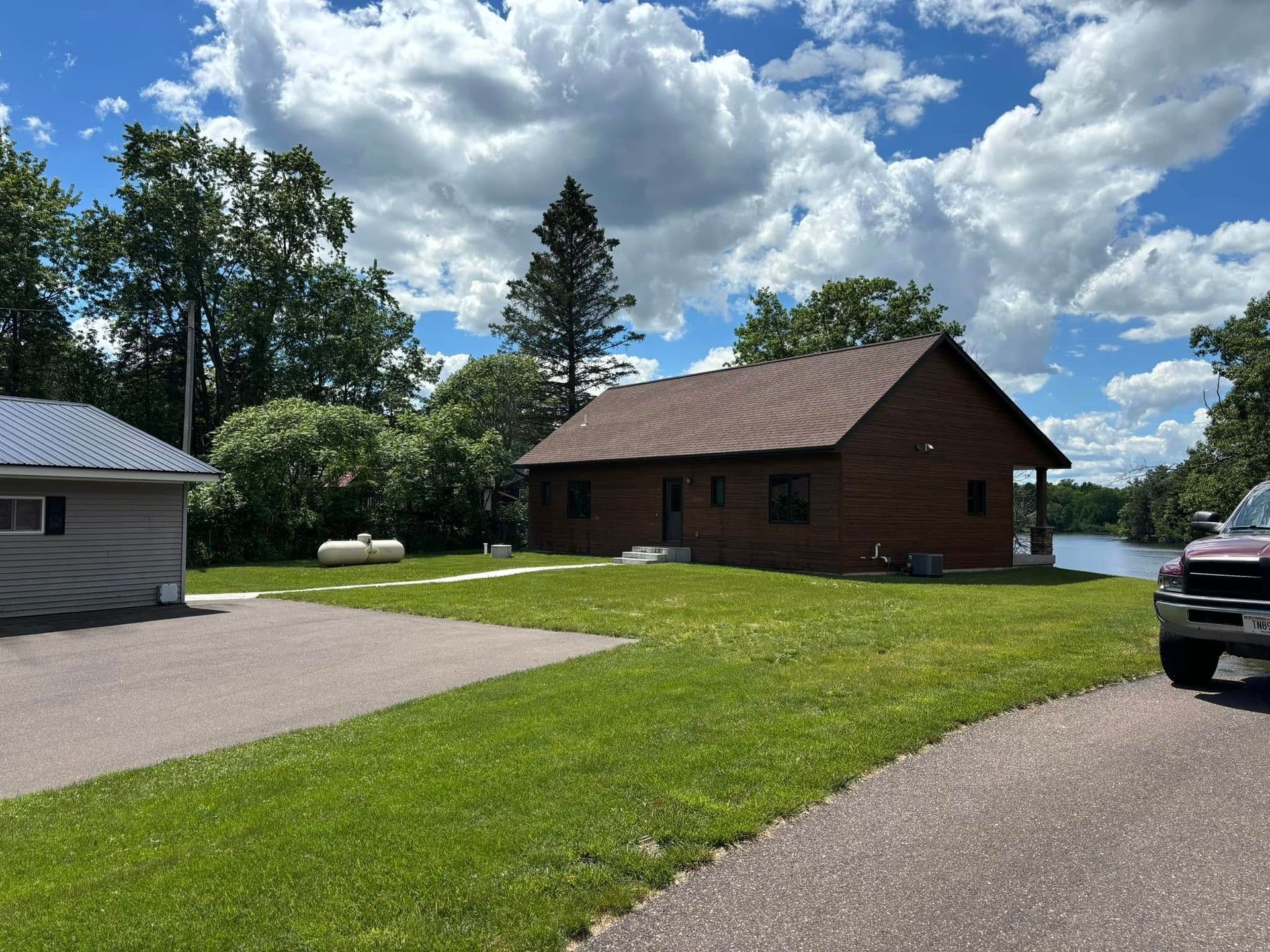 Rural cabin with brown roof beside a paved driveway, green lawn, and cloudy blue sky