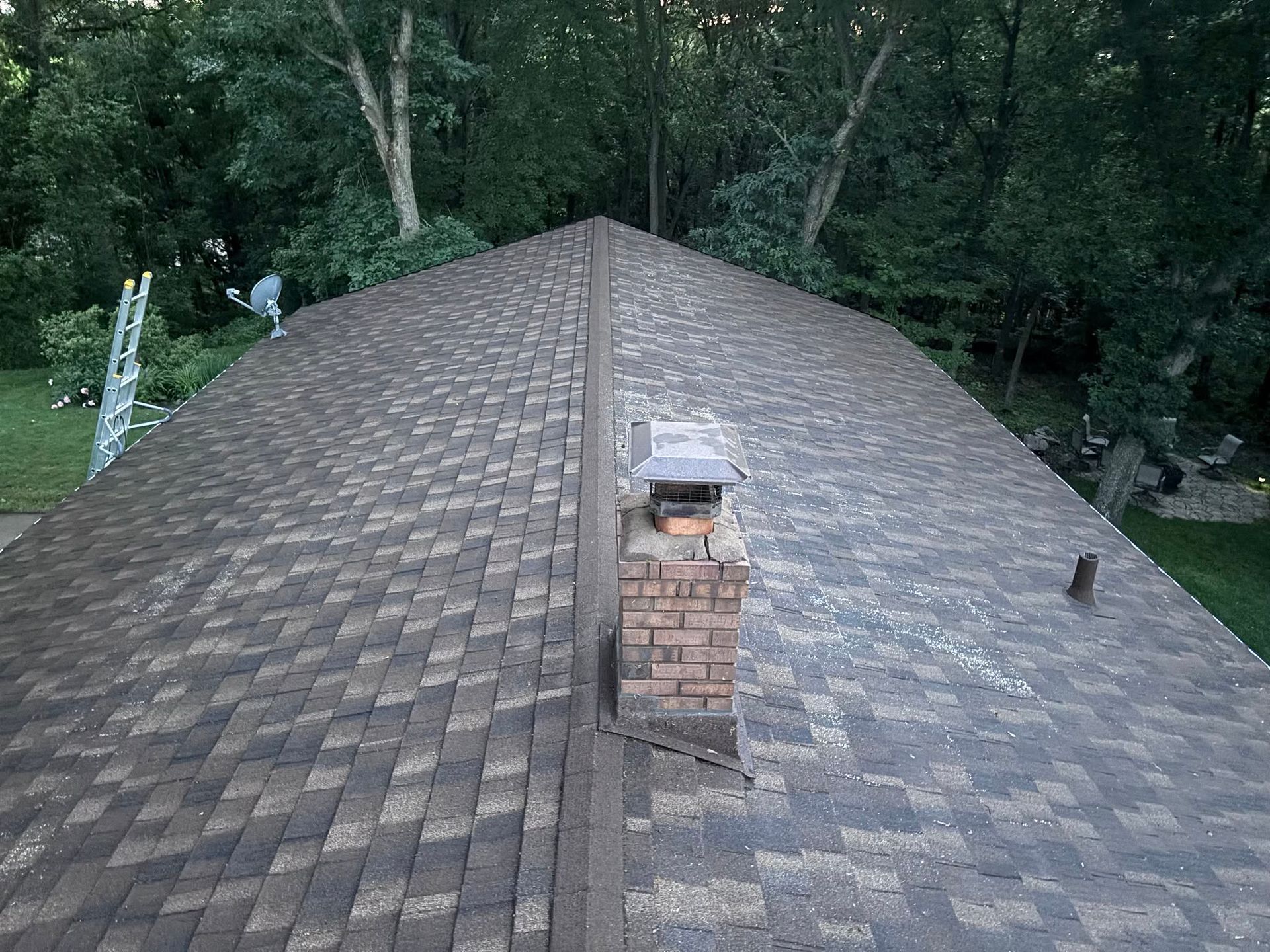 Shingled roof with a central brick chimney, surrounded by trees.