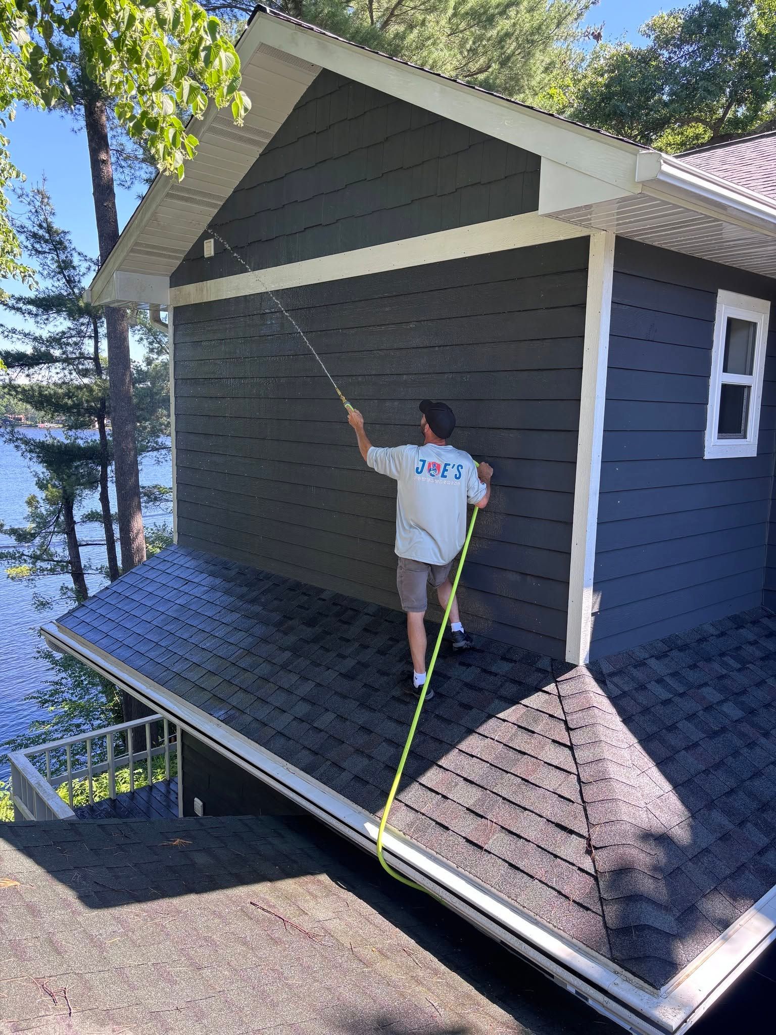 Person painting the exterior of a dark gray house on a rooftop deck with a long-handled roller.