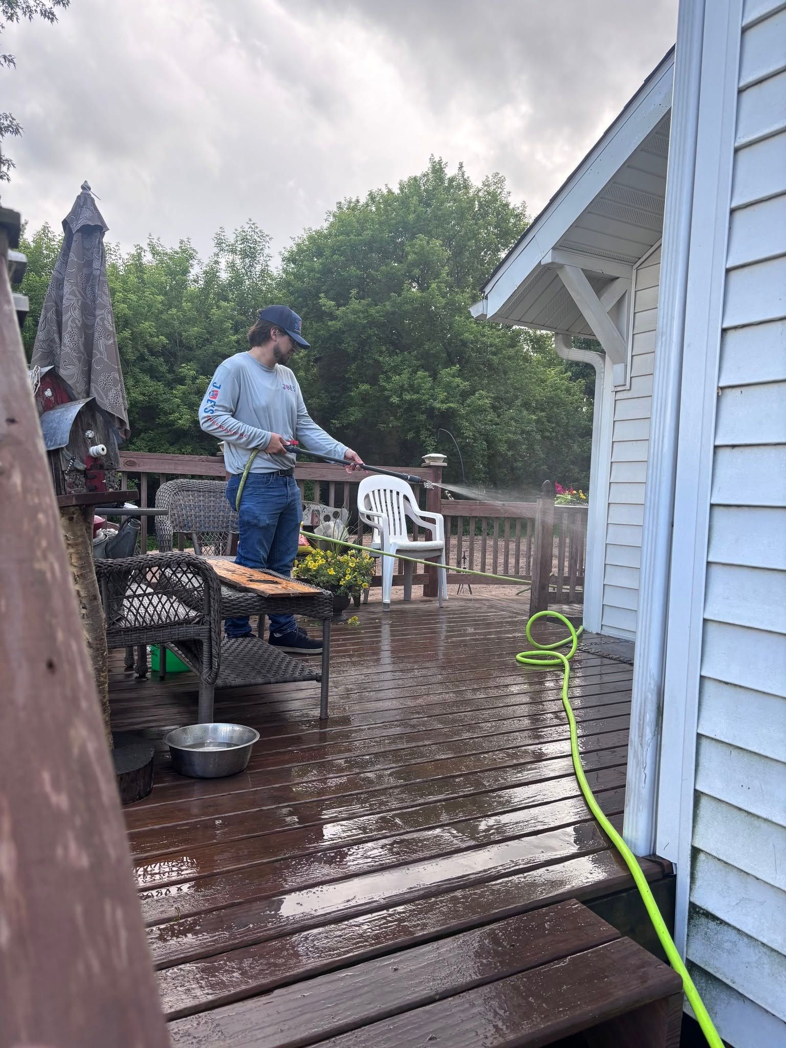 Person power-washing a wet wooden deck beside a blue house on a rainy day