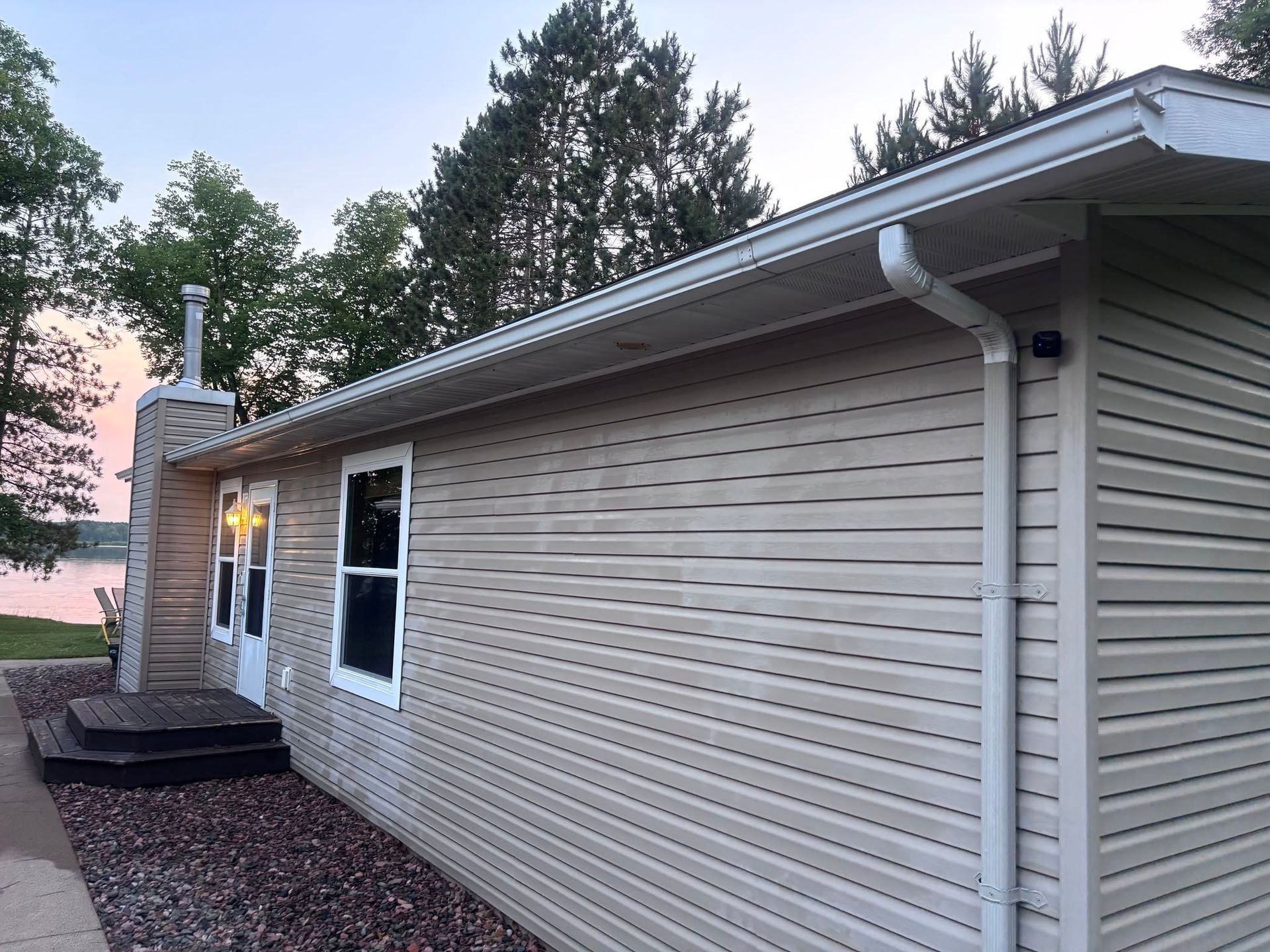 Single-story beige house exterior with horizontal siding, a lit porch, and trees at dusk