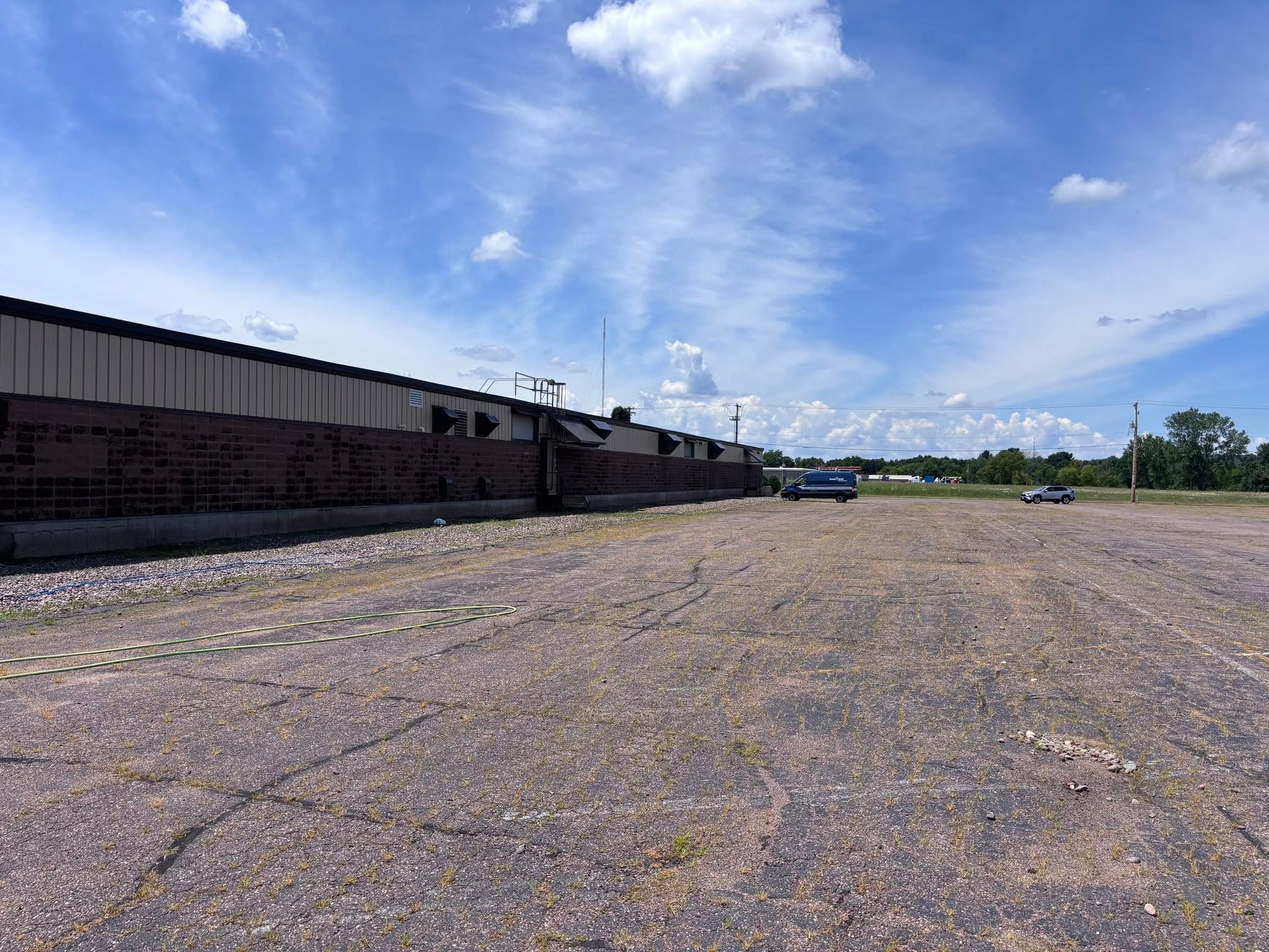 Empty gravel lot beside a long industrial building under a bright blue, partly cloudy sky