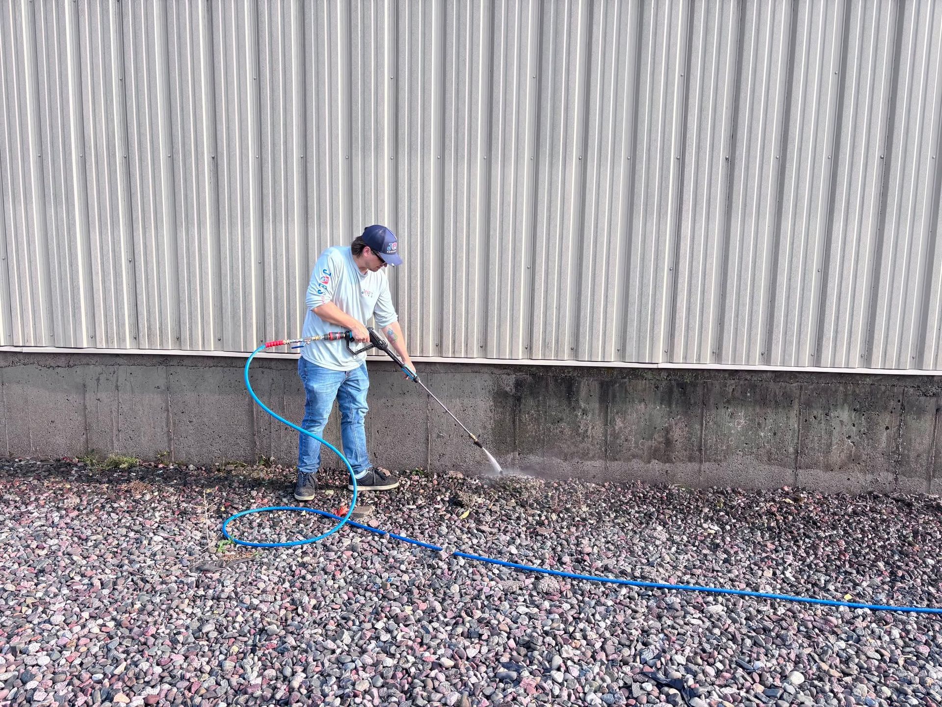 Person spraying water on rocky ground beside a corrugated metal wall with a blue hose