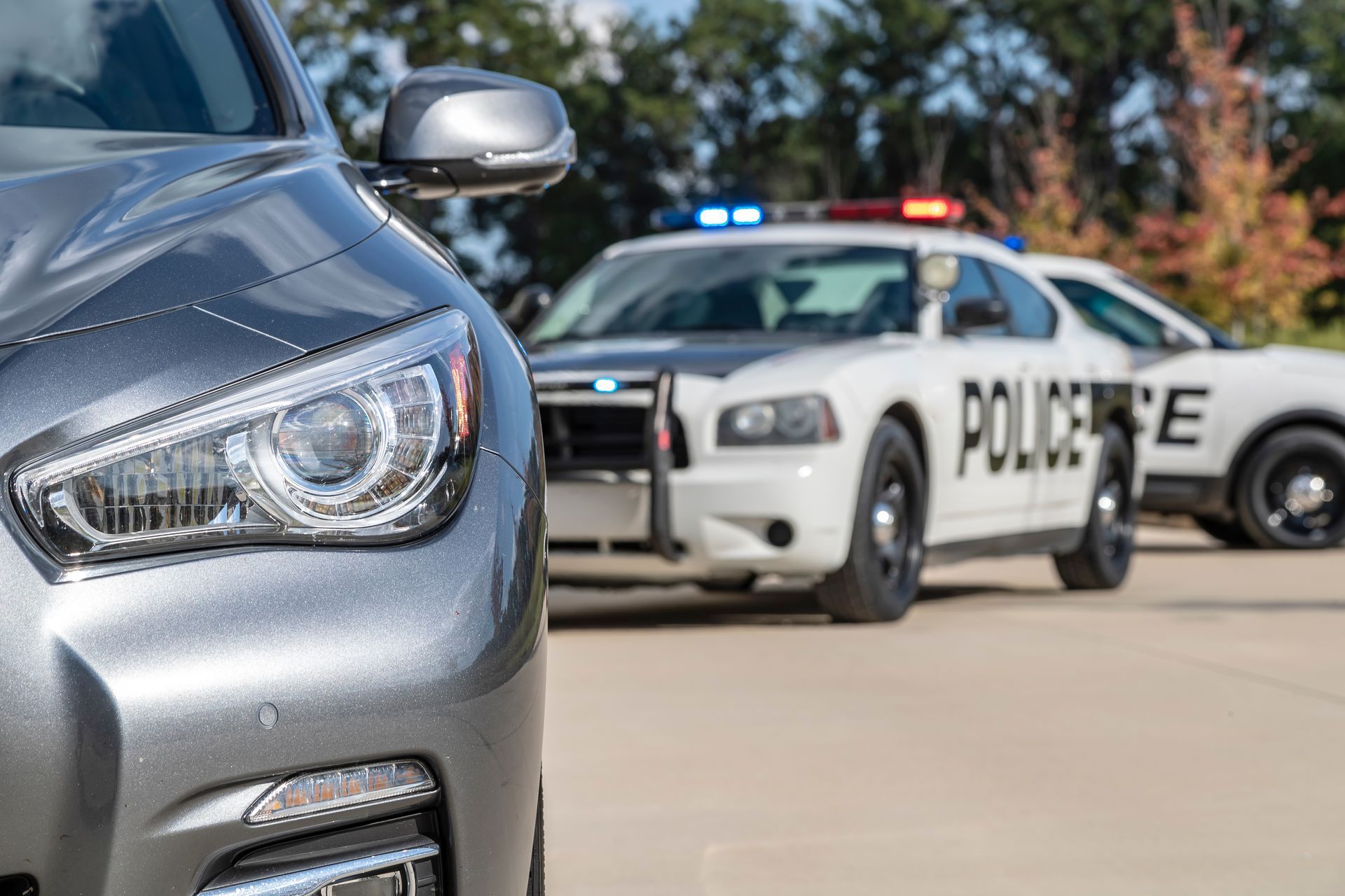 Gray car with front visible, police car in background with flashing lights.