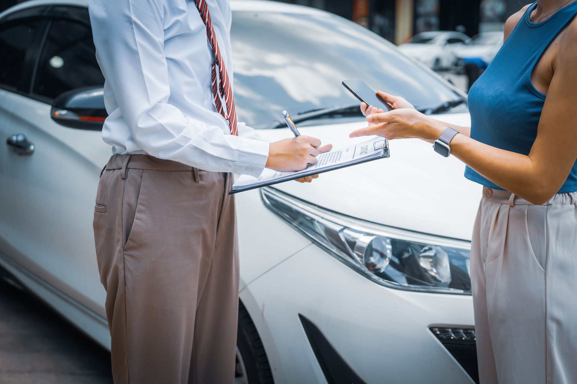 An agent with a clipboard discusses auto insurance coverage with a car owner beside a white sedan.