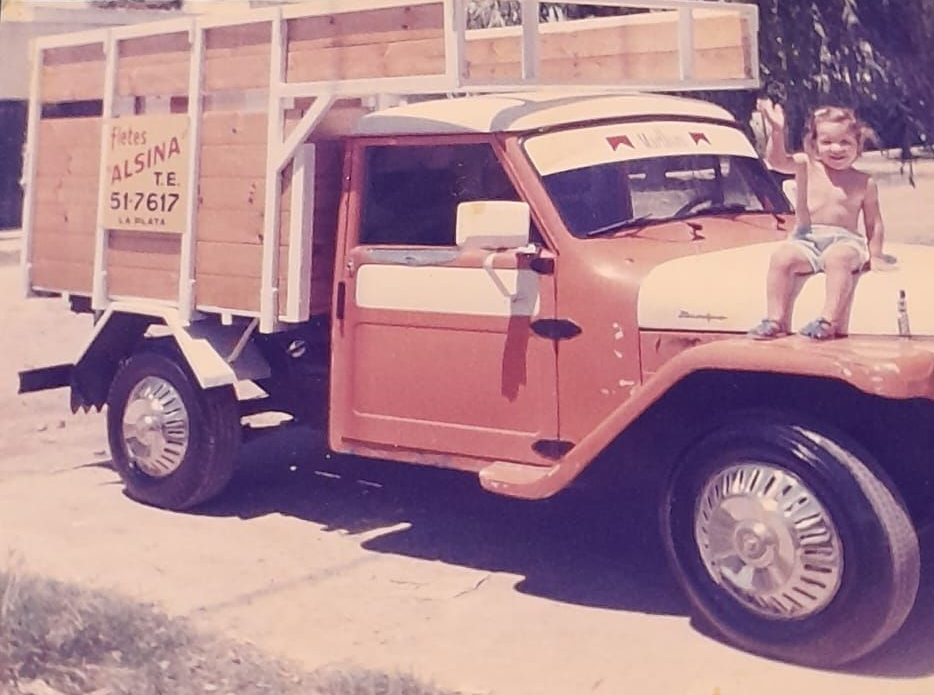 Camioneta pickup naranja antigua con caja de carga de madera, estacionada al aire libre con un niño sentado en el capó.