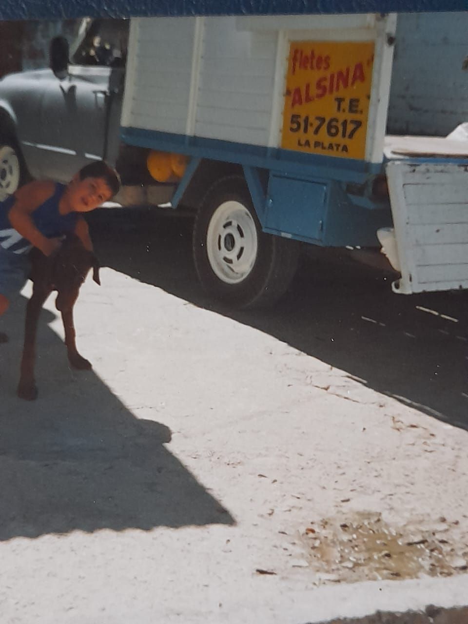 Un niño se agacha cerca de un camión azul y blanco con un letrero amarillo en el lateral, en una calle soleada.