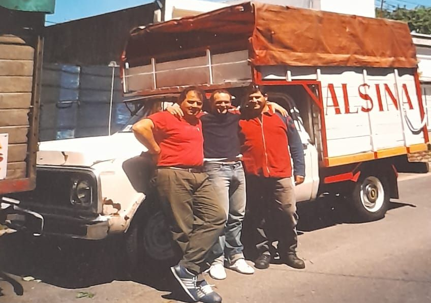 Tres personas con camisetas rojas posando junto a un camión de mudanzas blanco y rojo en una calle soleada.