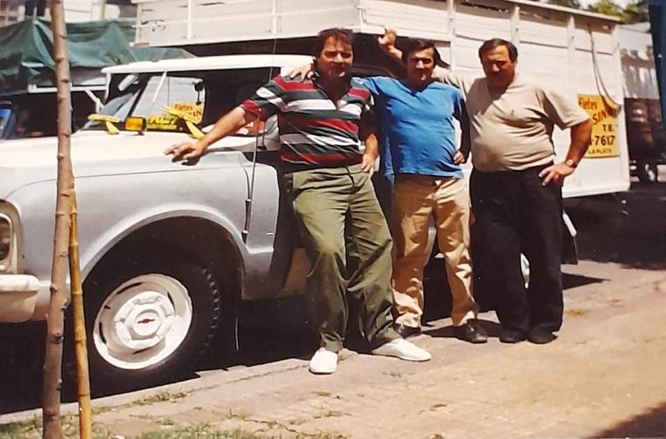 Tres personas de pie junto a una camioneta plateada en una calle soleada.