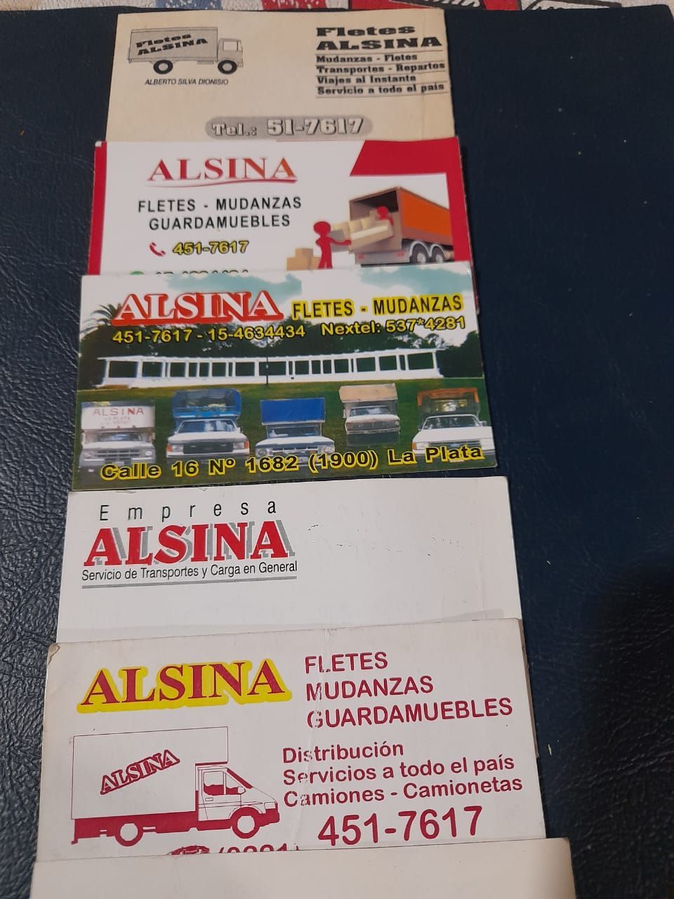 Pila de letreros y folletos comerciales de ALSINA con logotipos rojos y amarillos sobre una superficie oscura.