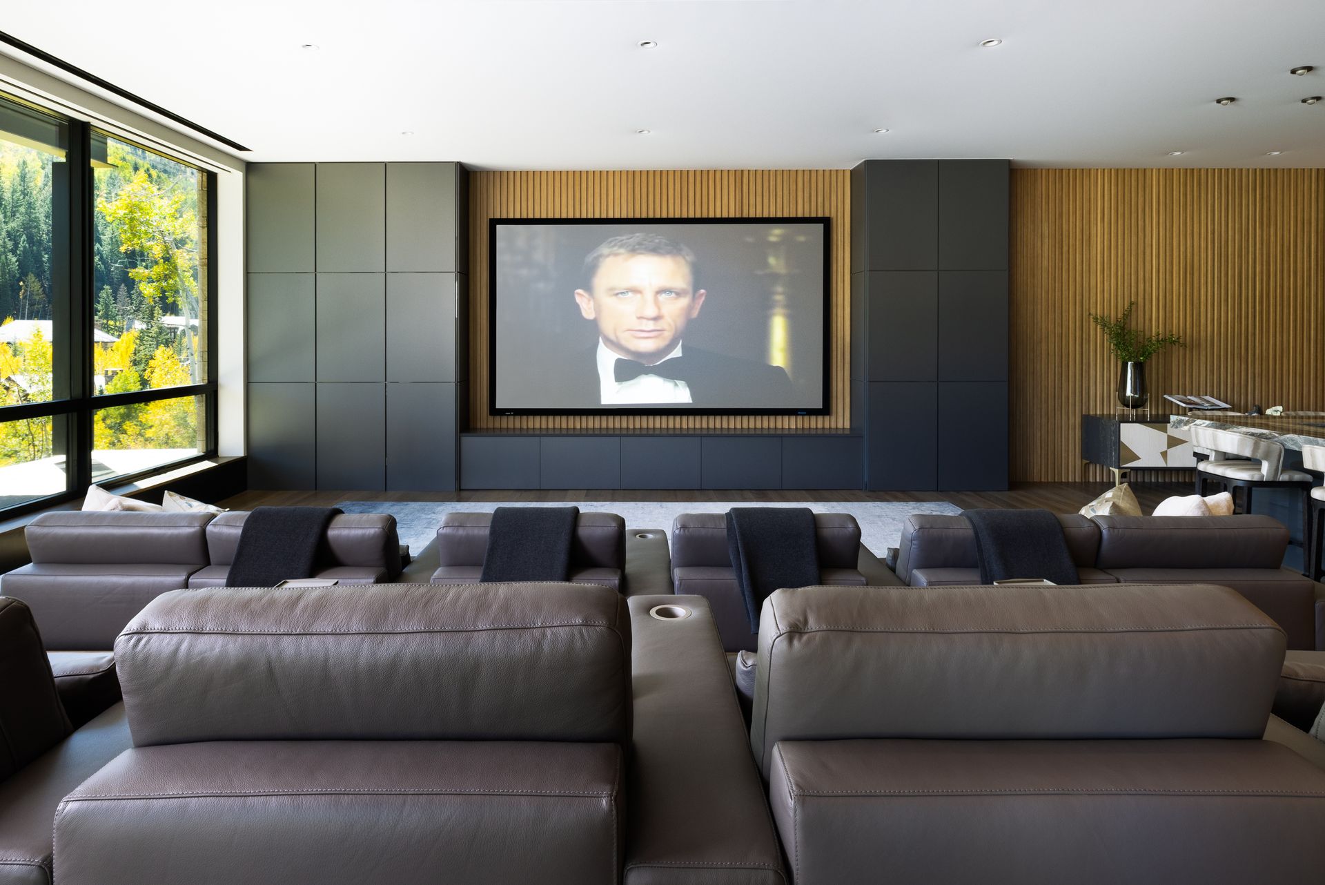 A living room with a large screen on the wall and a man in a tuxedo on it.