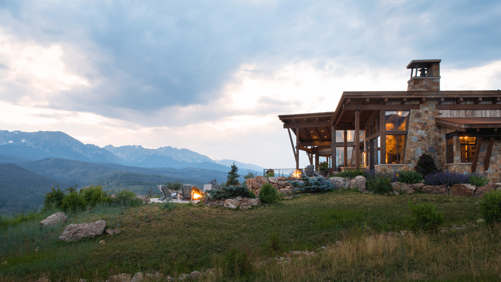 A large house is sitting on top of a grassy hill with mountains in the background.