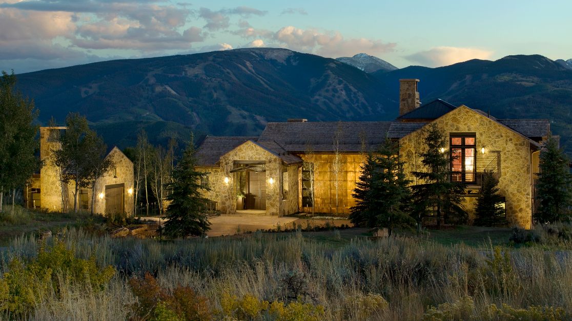A large house is sitting in the middle of a field with mountains in the background.
