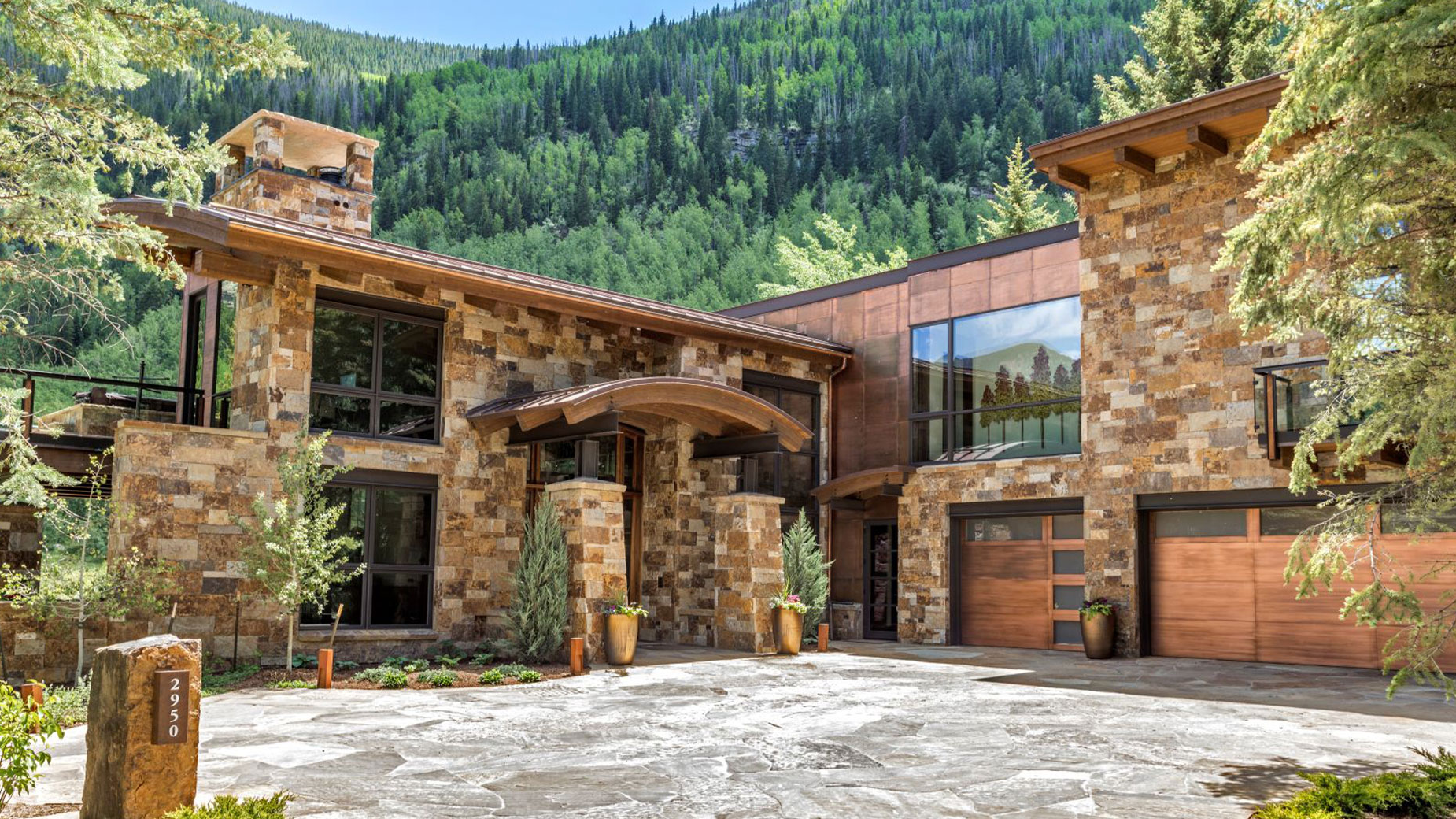 A large stone house with a lot of windows is surrounded by trees and mountains.