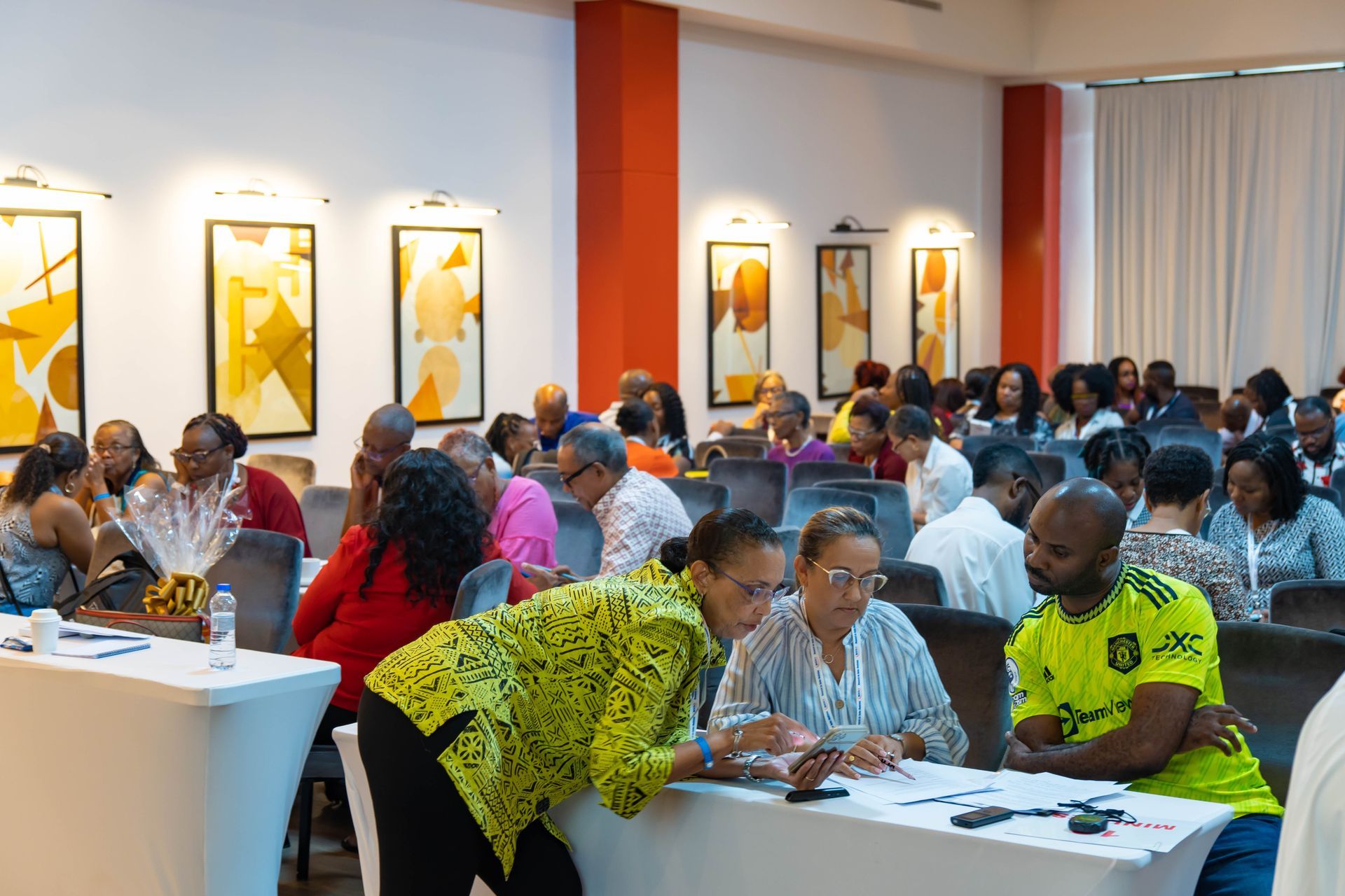 People seated at tables in a bright conference room, listening and taking notes.