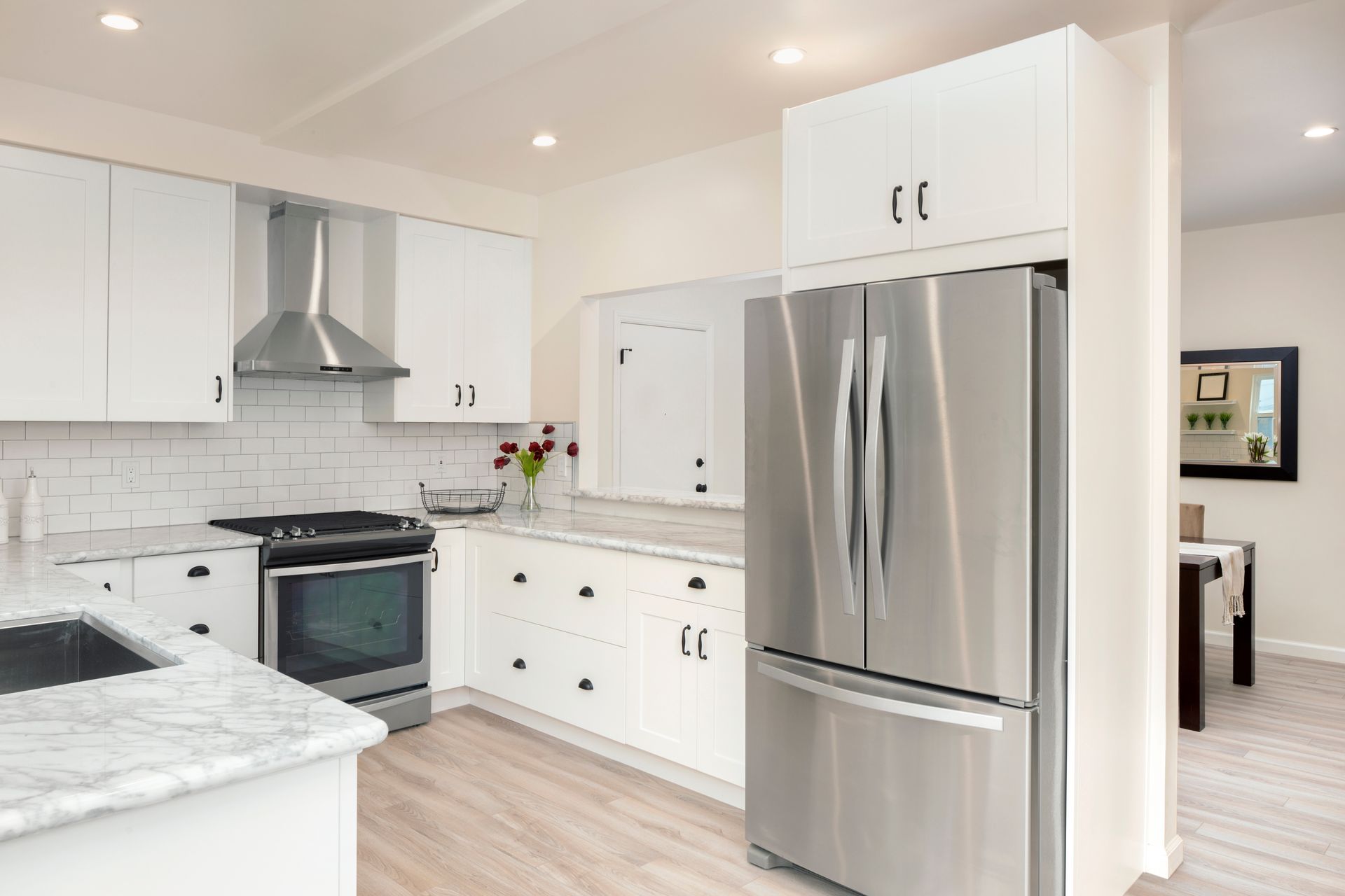 Bright modern white kitchen with stainless steel refrigerator and stove, marble countertops, and light wood floor